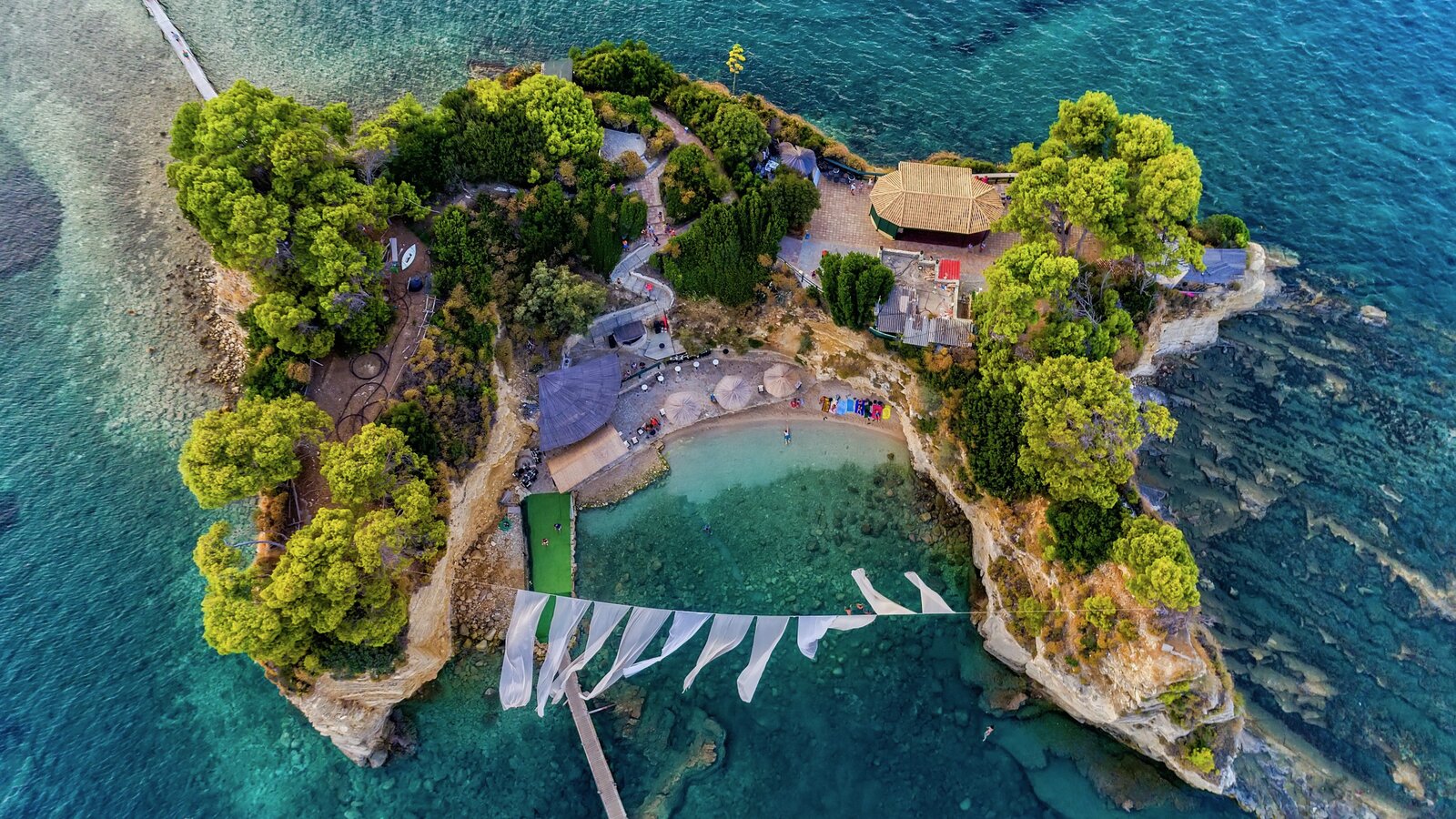 Aerial view of Cameo Island, a tiny tree-covered islet in turquoise water connected by a long wooden footbridge to the Zakynthos coast