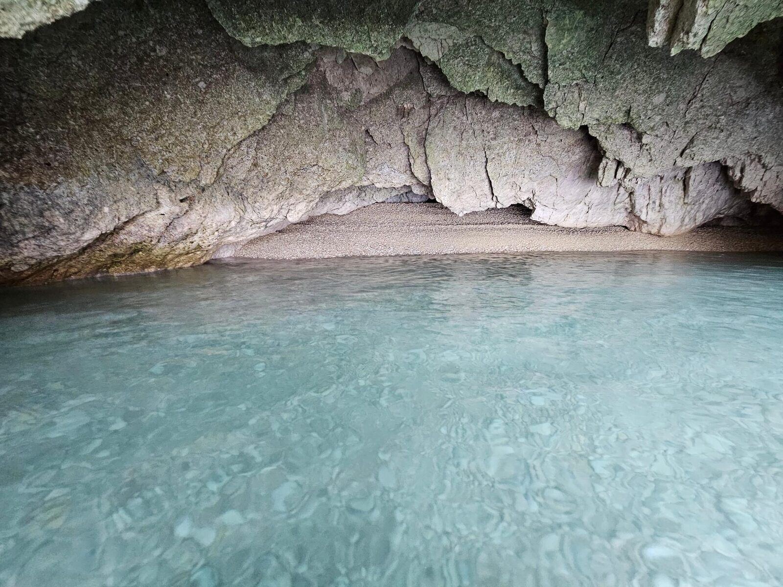 Water-level view inside a sea cave with a rocky overhang and pebble beach beyond