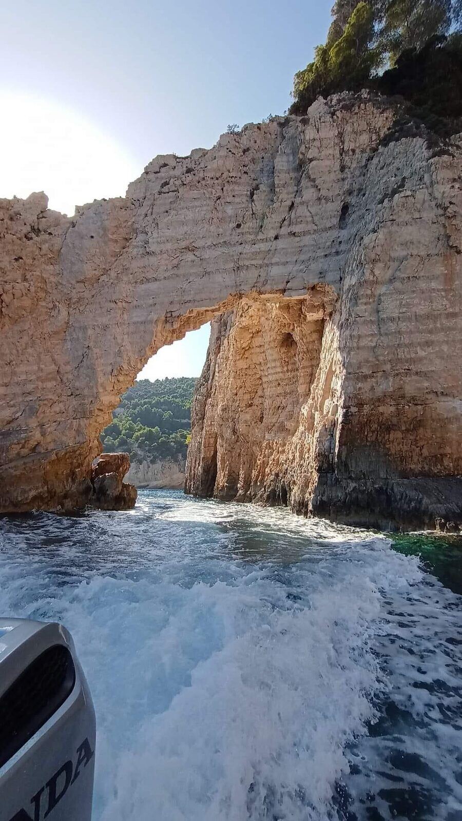 Speed boat wake passing through a towering limestone sea arch at the Keri Caves