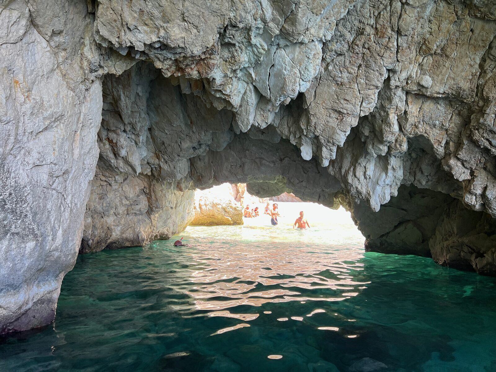 Inside a large Keri sea cave looking out through the opening to swimmers on a sun-lit beach