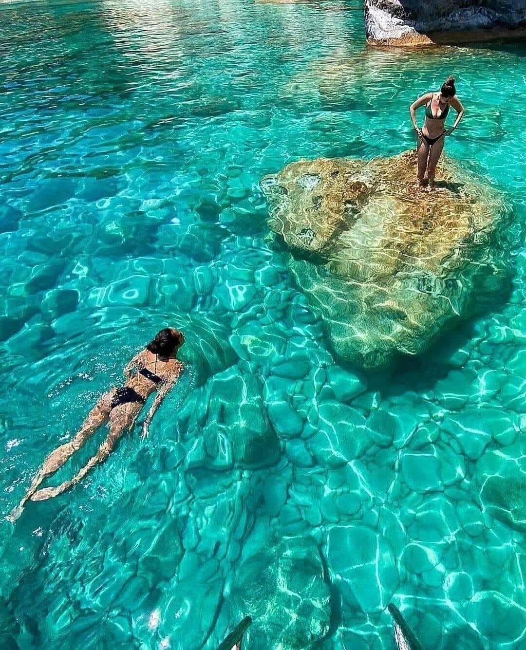 Two swimmers in crystal turquoise water beside a submerged rock at the Keri Caves
