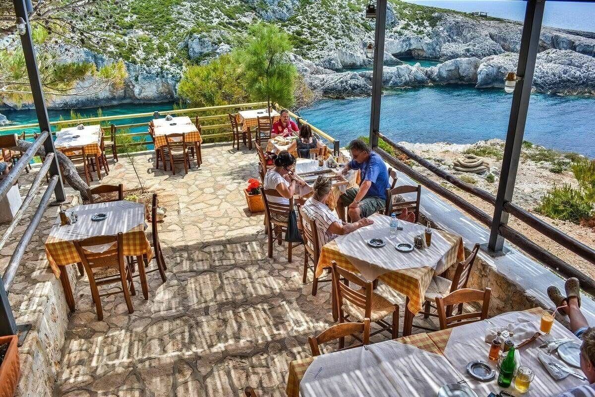 Inside the Porto Limnionas taverna with diners at chequered tablecloths and the rocky coastline behind